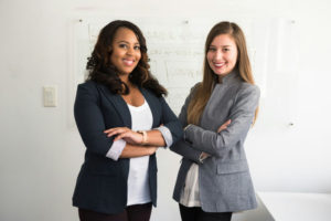 two-women-in-suit-standing-smiling-with-their-hands-folded