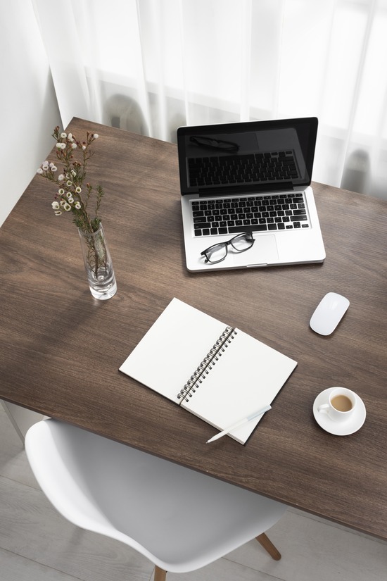 Brightly lit room with white curtains containing a wooden table and white chair, a flower vase, coffee cup, notebook, mouse, and glasses