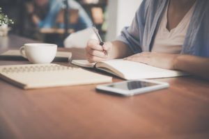 woman-writing-on-a-notebook-beside-tea-cup-and-tablet-computer