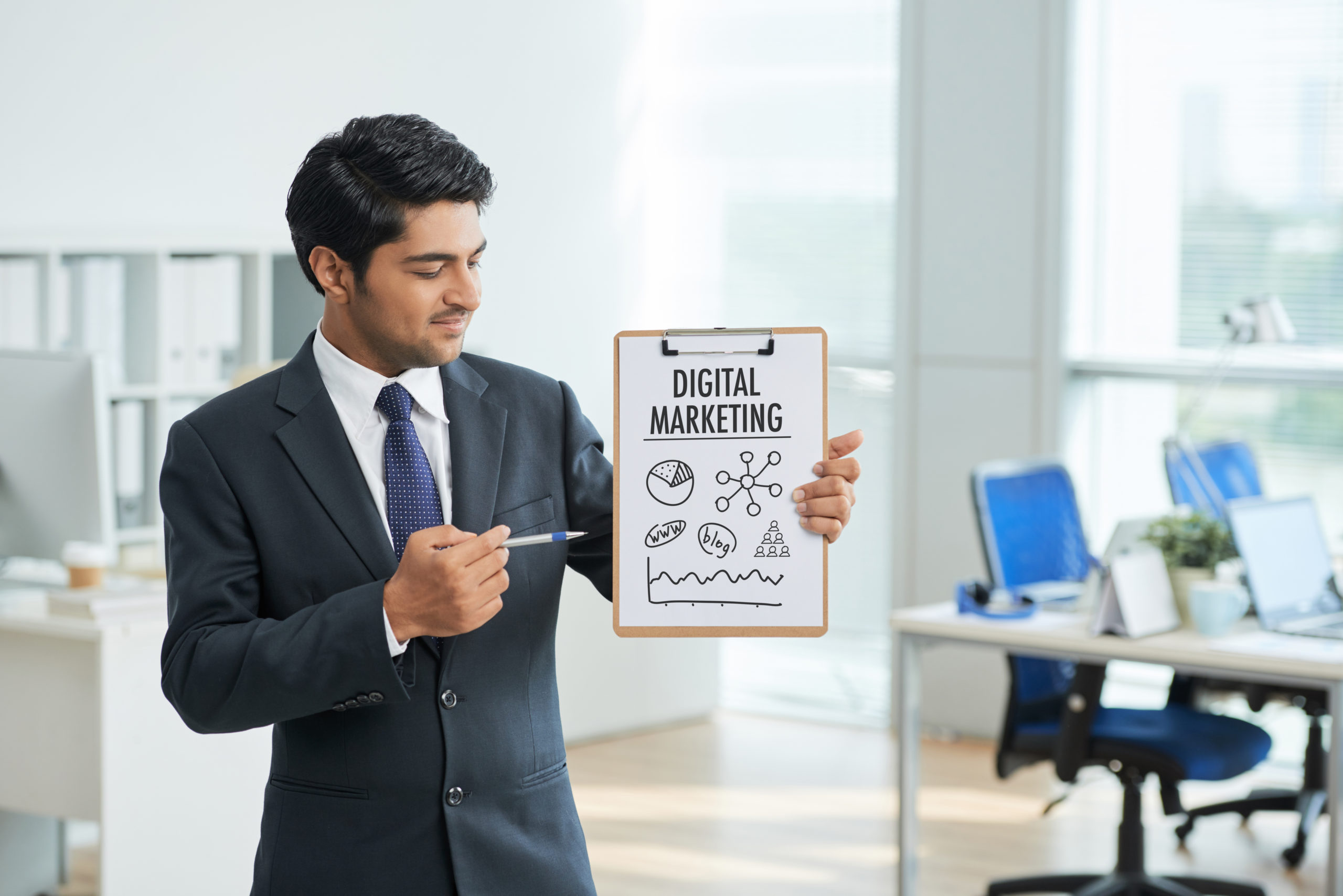 Man in suit standing in office with clipboard and pointing to poster with words digital marketing outsourcing