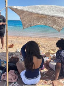 muqadsa-sitting -under-a-shade-by-the beach with friends, the-sea-stretching-out-in-front-of-them