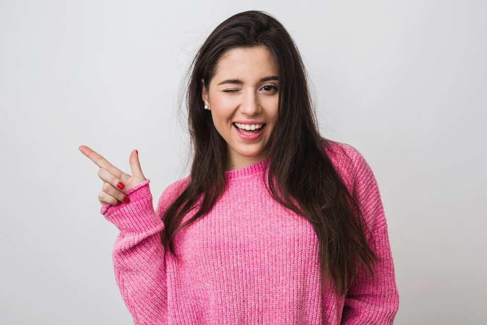 young-beautiful-woman-pink-warm-sweater-natural-look-smiling-pointing-finger-aside-winking-portrait-isolated-long-hair-funny-face-expression-positive-mood