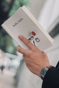 Close-up of a hand holding a book featuring a coffee cup and clock illustration, representing productivity and daily routines.