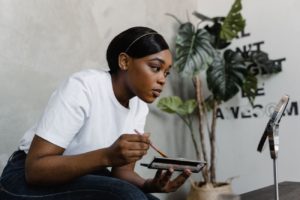 A melanin woman examining her skin in a mirror, reflecting the beauty industry's habit of manufacturing flaws before selling solutions