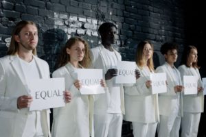 A diverse group of six people in white outfits holding equality signs standing together against a dark wall.