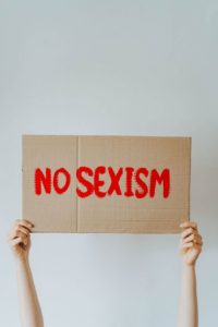 A person holds a cardboard sign reading “NO SEXISM” in bold red letters against a plain light background.