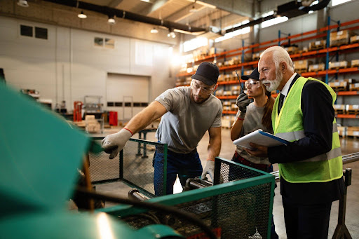 Two male workers in an industrial workshop examine a piece of machinery together. A younger worker in a gray T-shirt gestures toward the equipment while an older worker wearing a high-visibility vest holds a clipboard and pen and observes the process. The image illustrates hands-on skill development, practical experience, and knowledge sharing in the workplace.