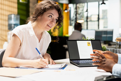 A recruiter woman with curly hair sits at a desk, writing notes in a notebook beside an open laptop while discussing information with a candidate seated across from her.